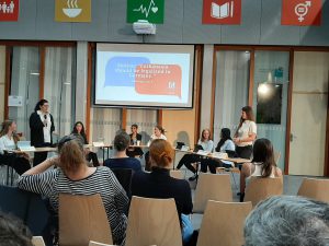 Eine Diskussionsveranstaltung in einem modernen Raum mit mehreren Teilnehmerinnen. Im Hintergrund ist eine Präsentation auf einem Bildschirm zu sehen, die Fragen stellt. Die Zuschauer sitzen in Stühlen und schauen auf die Bühne, wo eine Frau spricht. An den Wänden hängen verschiedene Symbole, die verschiedene Themen darstellen.