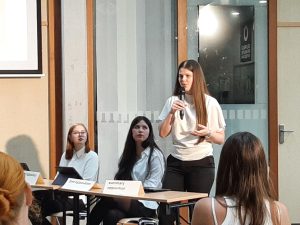 Eine Frau steht vor einem Tisch und spricht in ein Mikrofon, während zwei andere Frauen auf Stühlen sitzen. Alle drei Frauen tragen weiße Blusen. Die Frau am Tisch hat ein Notizbuch oder ein Tablet vor sich. Im Hintergrund sind Fenster und eine Präsentation sichtbar. Die Szene findet in einem hellen Raum statt, der für eine Präsentation oder ein Meeting genutzt wird.