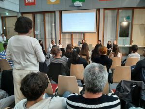Eine Gruppe von Menschen sitzt in einem Raum, während eine Diskussion oder Präsentation stattfindet. Im Vordergrund sind Teilnehmer zu sehen, die auf die Redner blicken. An der Wand ist ein Bildschirm mit Text zu sehen. Die Atmosphäre wirkt informell und interaktiv, mit Stühlen in Reihen und einer offenen Sitzanordnung.