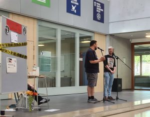 Zwei Personen stehen auf einer Bühne in einem hellen Raum und sprechen miteinander. Eine Person hält ein Mikrofon und spricht, während die andere zuhört und lächelt. Im Hintergrund sind Informationsschilder an der Wand zu sehen. Auf dem Boden steht ein kleiner Tisch mit einer Blumenvase und weiteren Gegenständen.