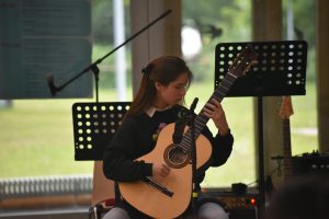 Eine junge Frau spielt konzentriert auf einer akustischen Gitarre. Sie sitzt auf einem Stuhl und trägt ein dunkles Sweatshirt. Im Hintergrund sind Musikständer mit Noten zu sehen, und ein Teil der Umgebung ist grün und hell erleuchtet.