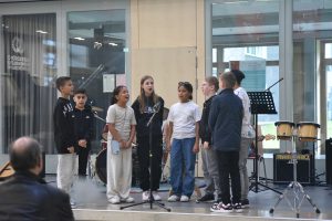 Eine Gruppe von sechs Kindern steht auf einer Bühne und singt in ein Mikrofon. Sie tragen legere Kleidung und scheinen konzentriert bei der Darbietung zu sein. Im Hintergrund sind Musikinstrumente und Notenständer zu sehen. Zuschauer sitzen in der Nähe und hören aufmerksam zu.