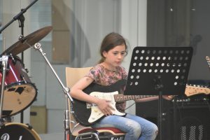 Ein Mädchen sitzt auf einem Stuhl und spielt eine E-Gitarre. Sie trägt ein buntes T-Shirt und hat lange, braune Haare. Vor ihr steht ein Notenständer mit Notenblättern. Im Hintergrund sind Schlagzeug und weitere Musikinstrumente sichtbar. Die Umgebung wirkt hell und einladend.