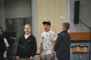 Drei Kinder stehen auf einer Bühne und singen zusammen. Einer der Jungen trägt ein T-Shirt mit bunten Motiven, ein anderer hat eine Mütze auf, und der dritte Junge ist in einem dunkelblauen Pullover gekleidet. Im Hintergrund sind Musikinstrumente und Mikrofone zu sehen.
