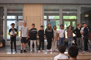 Eine Gruppe von zehn Kindern steht auf einer Bühne in einem Innenraum. Sie sind in verschiedenen Freizeitkleidungen gekleidet und schauen in Richtung des Publikums. Im Hintergrund sind Fenster und eine Musikanlage zu sehen.