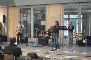 Eine Band spielt live auf einer Bühne in einem modernen Raum. Ein Sänger steht in der Mitte, während ein Gitarrist, ein Schlagzeuger und ein Geiger sie begleiten. Im Vordergrund sind einige Zuschauer zu sehen, die der Aufführung aufmerksam zuhören.