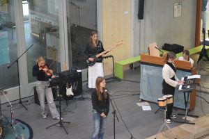 Eine Musikgruppe besteht aus fünf Mitgliedern, die auf einer Bühne auftreten. Eine Sängerin steht vorne, während im Hintergrund eine Musikerin am Keyboard spielt. Eine weitere Musikerin spielt E-Bass, und eine Violinistin ist ebenfalls zu sehen. Auf der linken Seite steht ein Schlagzeug. Die Umgebung ist hell und modern, mit großen Fenstern und einer offenen Raumgestaltung.