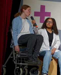 Zwei Personen auf einer Bühne in einem Theaterstück. Eine Person sitzt in einem Rollstuhl und hält ein Mikrofon, während die andere Person, die einen weißen Kittel trägt, interessiert zuhört. Im Hintergrund ist ein rotes Kreuz auf einer grauen Wand sichtbar.