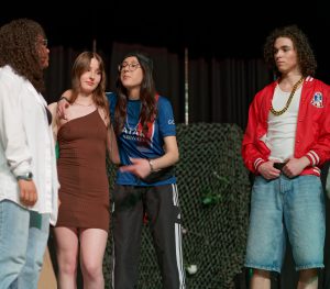 Vier junge Menschen stehen auf einer Bühne in einem Theater. Zwei von ihnen tragen sportliche Kleidung, während die anderen lässige Outfits anhaben. Die Gruppe scheint in ein Gespräch vertieft zu sein, und die Atmosphäre wirkt freundlich und lebhaft. Im Hintergrund sind einfache Bühnenrequisiten zu sehen.