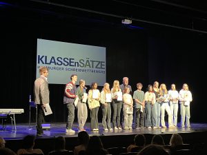 Eine Gruppe von Schülerinnen und Schülern steht auf einer Bühne bei einem Schreibwettbewerb. Im Hintergrund ist ein Banner mit dem Titel 