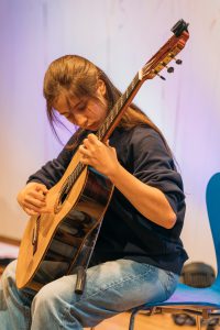 Eine junge Frau sitzt auf einem Stuhl und spielt konzentriert auf einer akustischen Gitarre. Sie hat lange, braune Haare und trägt ein dunkelblaues Oberteil sowie Jeans. Der Hintergrund ist hell und unscharf, mit sanften Farben, die eine entspannte Atmosphäre schaffen.
