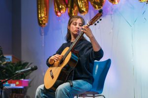 Ein junger Mensch sitzt auf einem Stuhl und spielt Gitarre. Im Hintergrund sind goldene Luftballons und eine festliche Dekoration zu sehen. Der Raum wirkt einladend und freundlich.