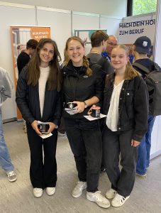 Drei junge Frauen stehen in einem Raum, lächeln und halten jeweils eine Tasse in der Hand. Sie tragen lässige Kleidung in dunklen Farbtönen. Im Hintergrund sind verschwommene Personen und Banner sichtbar, die auf eine Veranstaltung hinweisen.
