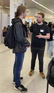 Zwei junge Männer stehen in einem modernen Raum und führen ein Gespräch. Der Mann links trägt einen schwarzen Rucksack und hat lange Haare, während der Mann rechts ein schwarzes T-Shirt und braune Schuhe trägt. Im Hintergrund sind weitere Personen und Stände zu sehen, die auf eine Veranstaltung hinweisen.
