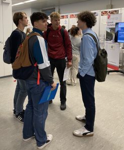 Vier junge Männer stehen in einem Raum und unterhalten sich. Sie tragen unterschiedliche Kleidung und Rucksäcke. Im Hintergrund sind Plakate und Informationsstände sichtbar, die auf eine Veranstaltung hinweisen.