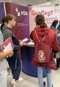 Zwei Jugendliche stehen an einem Messestand mit dem Logo der HSB (Hochschule Bremen). Der Junge links hält Informationsmaterial in der Hand, während das Mädchen rechts mit einem Rucksack steht und ebenfalls interessiert scheint. Im Hintergrund ist ein weiteres Banner sichtbar, das das Thema 