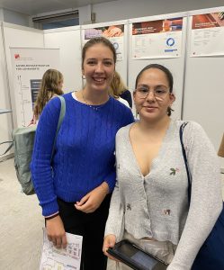 Zwei junge Frauen stehen lächelnd nebeneinander vor einem Hintergrund mit Informationsständen. Die Frau links trägt einen blauen Pullover und hat lange braune Haare. Die Frau rechts trägt einen hellen Pullover mit V-Ausschnitt und hat eine Brille. Beide halten Unterlagen in den Händen.