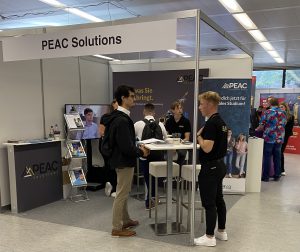 Messestand von PEAC Solutions auf einer Veranstaltung. Im Vordergrund stehen Personen, die miteinander sprechen und Informationsmaterial austauschen. Im Hintergrund sind Plakate und Displays zu sehen. Der Stand ist hell beleuchtet und wirkt einladend.