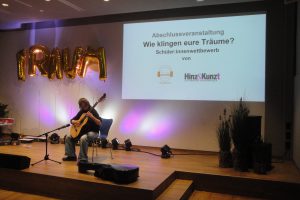 Ein Musiker sitzt mit einer Gitarre auf einer Bühne. Im Hintergrund ist eine Leinwand mit der Aufschrift 