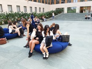 Eine Gruppe von Frauen sitzt auf blauen, runden Sitzmöbeln in einem Innenhof. Im Hintergrund sind Pflanzen und moderne Architektur zu sehen. Einige Frauen unterhalten sich, während andere auf ihren Handys sind. Der Boden ist mit großen Fliesen gestaltet.