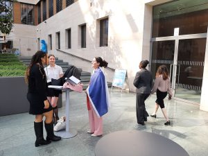 Fünf Personen stehen im Freien in einem modernen, lichtdurchfluteten Bereich vor einem Gebäude. Eine Frau mit langen, dunklen Haaren und schwarzen Stiefeln redet mit einer anderen Frau, die einen weißen Blazer trägt. Diese hat einen blauen Umhang an und steht an einem Stehtisch mit Unterlagen. Zwei weitere Frauen gehen im Hintergrund vorbei, während ein Mann in einem grauen Anzug ebenfalls in die Richtung des Eingangs geht. Der Boden ist aus Stein und es gibt einige Pflanzen im Hintergrund.