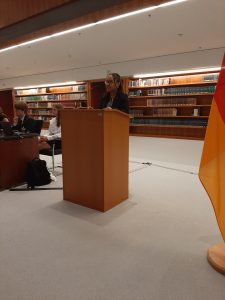 Eine Frau steht an einem Rednerpult in einem Raum mit Bücherregalen im Hintergrund. Sie trägt einen schwarzen Anzug und spricht, während eine deutsche Flagge an der rechten Seite des Bildes sichtbar ist. Im Hintergrund sind Personen an einem Tisch zu sehen. Der Raum ist gut beleuchtet und hat einen modernen, professionellen Eindruck.