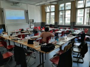 Ein Klassenraum mit mehreren Tischen und roten Stühlen. Auf den Tischen liegen verschiedene Materialien und persönliche Gegenstände. Im Hintergrund befindet sich ein Projektor, der auf eine Wand zeigt, und große Fenster lassen Tageslicht herein. Eine Person sitzt mit dem Rücken zur Kamera und schaut auf den Projektor.