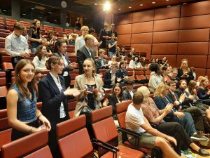 Eine Gruppe von Menschen sitzt und steht in einem Saal mit roten Sitzreihen. Einige der Anwesenden klatschen, während andere auf ihre Smartphones schauen oder sich unterhalten. Der Raum scheint gut besucht zu sein, mit einer Mischung aus formeller und informeller Kleidung. Im Hintergrund sind weitere Personen zu sehen, die sich in einer geselligen Atmosphäre aufhalten.
