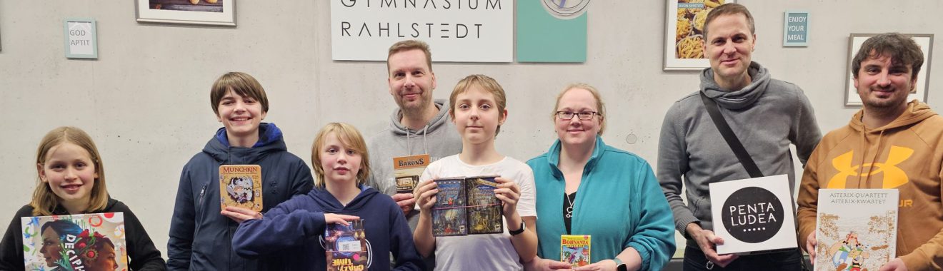 Gruppe von sieben Personen steht lächelnd vor einer Wand mit Kunstwerken. Sie halten verschiedene Bücher in den Händen. Im Hintergrund sind einige Bilder zu sehen, sowie ein Schild mit dem Text "SPANNSTOWN FAMILIERT".