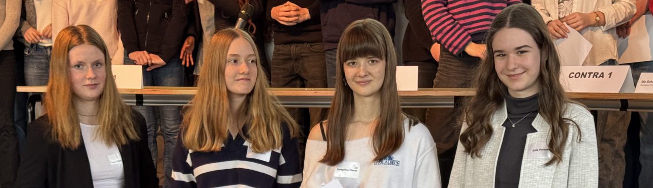 Vier junge Frauen stehen in einer Gruppe, lächeln und posieren für das Foto. Im Hintergrund sind weitere Personen sichtbar, die teilweise unscharf sind. Die Frauen tragen verschiedene Oberteile in unterschiedlichen Farben.