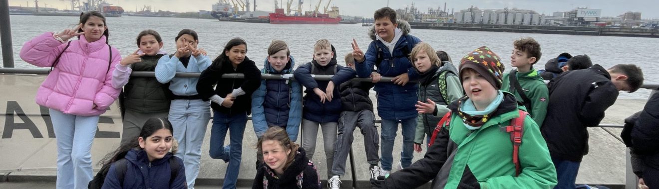 Gruppe von Jugendlichen in winterlicher Kleidung, die lächelnd und posierend am Ufer stehen. Einige sitzen auf dem Boden, während andere in einer Reihe stehen. Im Hintergrund ist ein Gewässer mit Schiffen und einem bewölkten Himmel zu sehen.