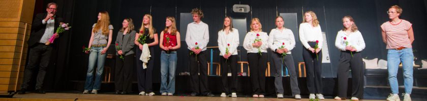 Eine Gruppe von zehn Personen steht auf einer Bühne. Sie sind in formeller Kleidung gekleidet und halten Blumen. Eine Person spricht, während die anderen aufmerksam zuhören. Im Hintergrund sind Vorhänge und einige Stühle sichtbar. Die Szene vermittelt eine feierliche Stimmung, möglicherweise im Rahmen einer Auszeichnung oder Zeremonie.