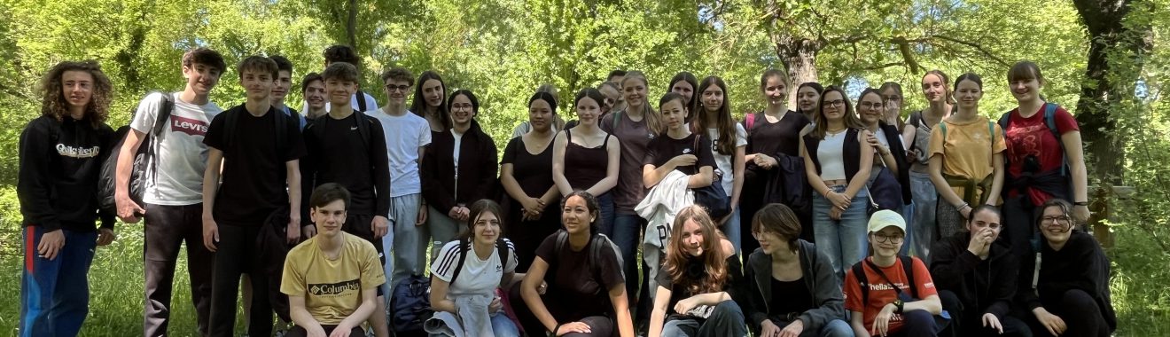 Gruppe von etwa 40 Personen, die auf einer Wiese im Freien steht, umgeben von Bäumen. Die Teilnehmer sind unterschiedlich gekleidet und posieren für das Foto. Es ist ein sonniger Tag mit blauem Himmel.