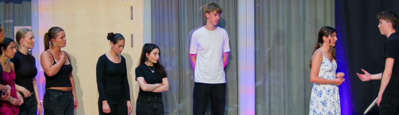 Eine Gruppe von sieben Personen steht auf einer Bühne. Einige der Darsteller tragen schwarze Kleidung, während andere farbige Outfits haben. Im Vordergrund führt eine Person ein Gespräch mit einer anderen, die ein weißes Kleid trägt. Im Hintergrund sind zwei Personen zu sehen, die in neutraler Kleidung stehen. Die Bühne ist mit einem neutralen Hintergrund und sanfter Beleuchtung gestaltet.