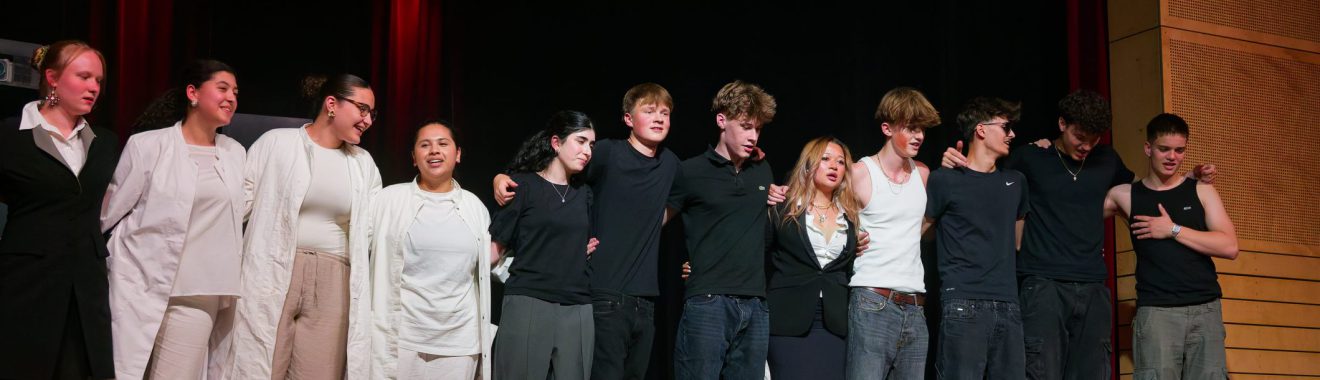 Gruppenfoto von Schauspielern auf der Bühne. Sie stehen nebeneinander und lächeln, einige tragen schwarze Kleidung, andere helle. Der Hintergrund ist dunkel, mit rotem Licht. Die Personen wirken stolz und glücklich, möglicherweise nach einer Aufführung.