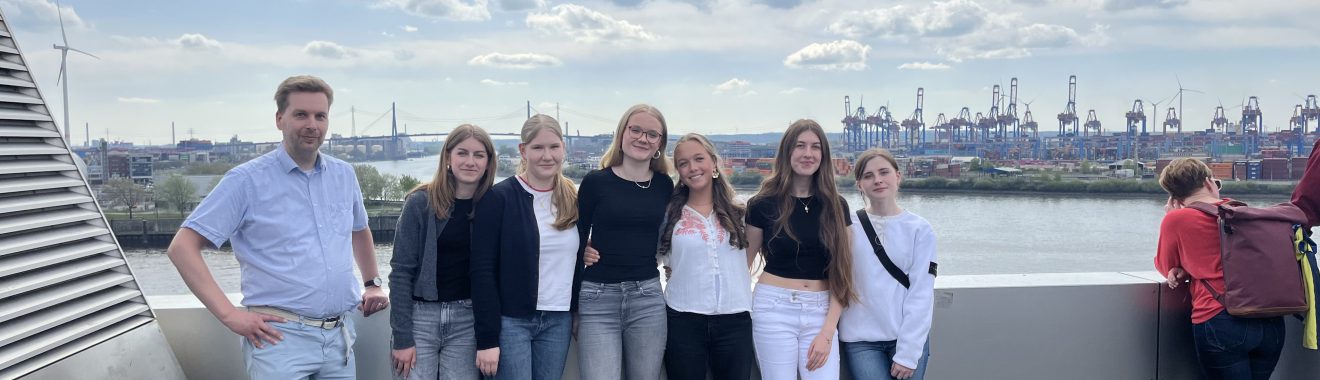 Eine Gruppe von sieben Personen steht auf einer Aussichtsplattform. Sie posieren lächelnd für das Foto, während im Hintergrund ein Fluss und eine Hafenlandschaft mit Kranen und Schiffen zu sehen sind. Der Himmel ist teilweise bewölkt.