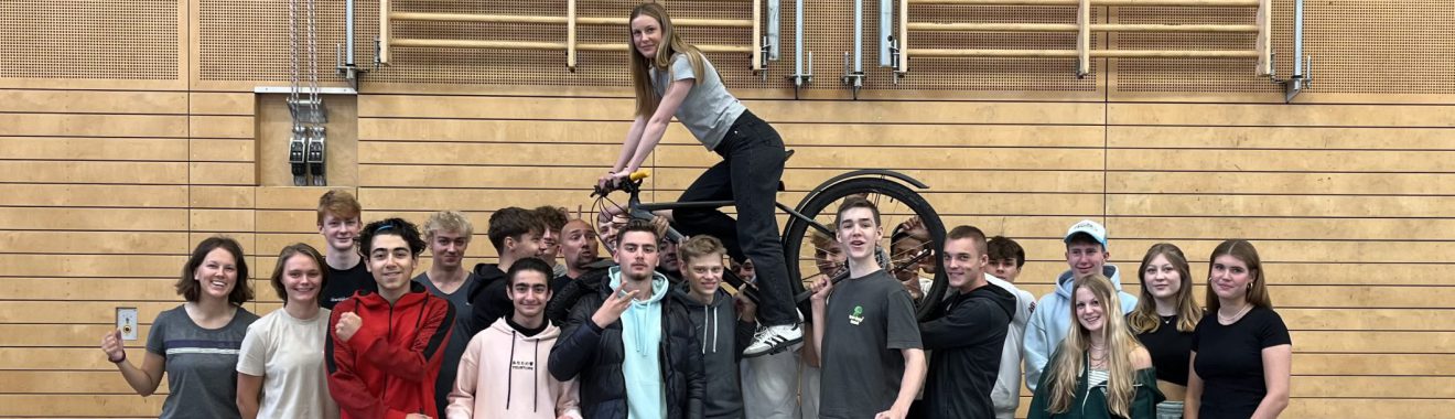 Gruppe von Jugendlichen, die in einer Halle zusammenstehen. Einige Personen posieren lächelnd, während eine Person auf einer anderen steht. Die Umgebung ist mit Holzpaneelen gestaltet, und im Hintergrund sind Sportgeräte sichtbar.