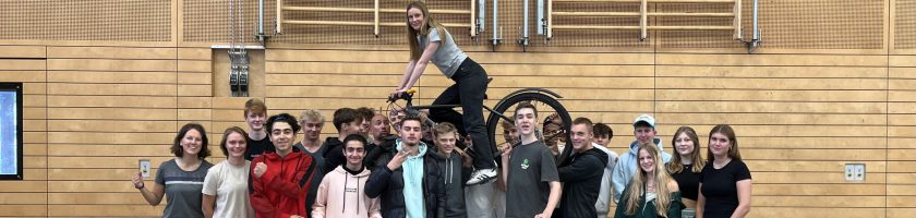 Gruppe von Jugendlichen, die in einer Halle zusammenstehen. Einige Personen posieren lächelnd, während eine Person auf einer anderen steht. Die Umgebung ist mit Holzpaneelen gestaltet, und im Hintergrund sind Sportgeräte sichtbar.