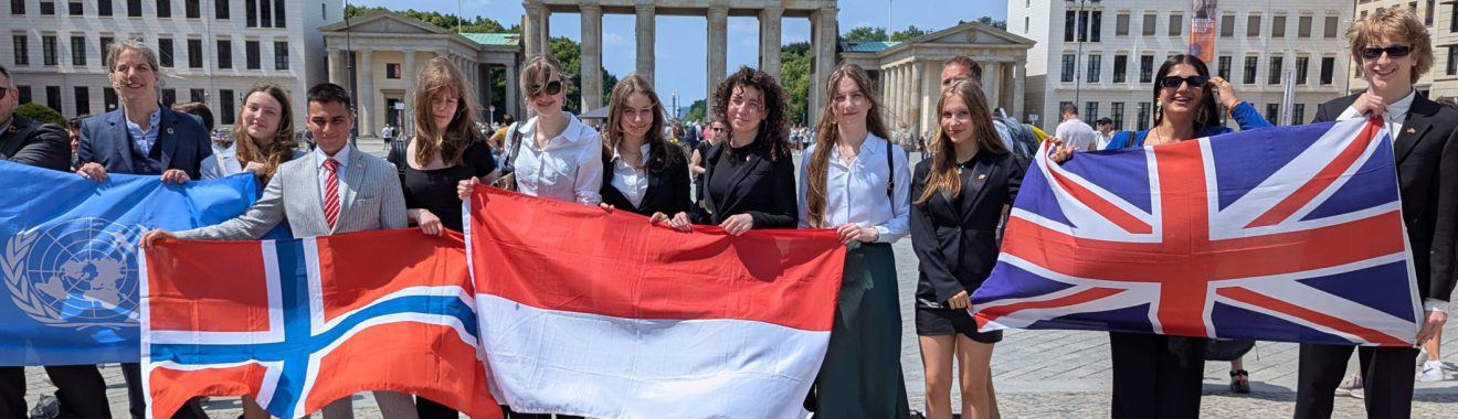 Gruppe von Menschen steht vor einem historischen Gebäude und hält verschiedene Nationalflaggen, darunter die Flaggen von Norwegen, Indonesien und Großbritannien. Es ist ein sonniger Tag mit klarem Himmel.