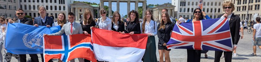 Gruppe von Menschen steht vor einem historischen Gebäude und hält verschiedene Nationalflaggen, darunter die Flaggen von Norwegen, Indonesien und Großbritannien. Es ist ein sonniger Tag mit klarem Himmel.