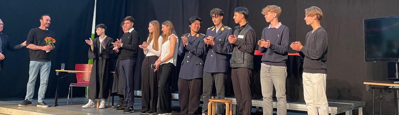 Eine Gruppe von acht Personen steht auf einer Bühne. Sie applaudieren und lächeln. Im Hintergrund sind ein Tisch und ein Bildschirm zu sehen. Die Personen tragen unterschiedliche Kleidung, darunter Schuluniformen und legere Outfits. Das Licht ist gedimmt, und die Atmosphäre wirkt festlich.