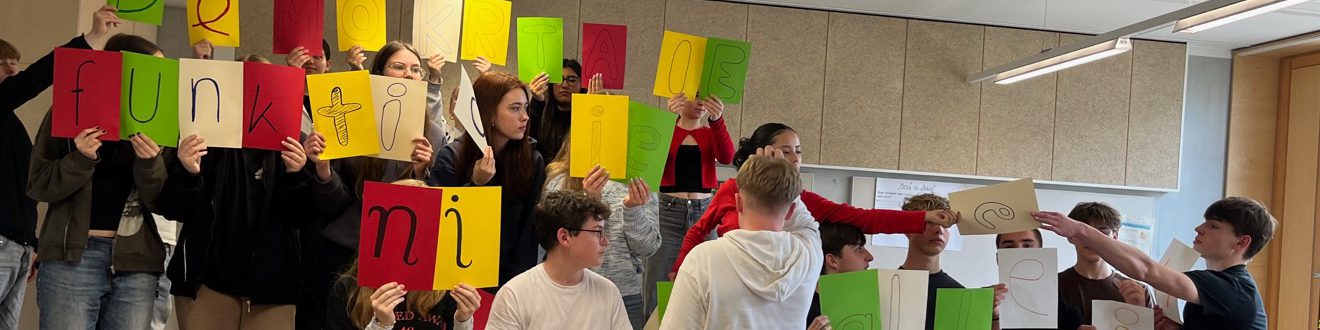 Eine Gruppe von Schülerinnen und Schülern steht in einem Raum und hält bunte Plakate in verschiedenen Farben hoch. Die Plakate zeigen Buchstaben, die zusammen ein Wort oder einen Satz bilden. Einige der Schülerinnen und Schüler sind aktiv miteinander beschäftigt, während andere in die Kamera blicken. Der Raum hat eine helle, freundliche Atmosphäre mit großen Fenstern.