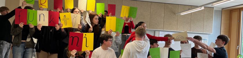 Eine Gruppe von Schülerinnen und Schülern steht in einem Raum und hält bunte Plakate in verschiedenen Farben hoch. Die Plakate zeigen Buchstaben, die zusammen ein Wort oder einen Satz bilden. Einige der Schülerinnen und Schüler sind aktiv miteinander beschäftigt, während andere in die Kamera blicken. Der Raum hat eine helle, freundliche Atmosphäre mit großen Fenstern.