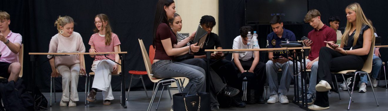 Eine Gruppe von Jugendlichen sitzt an Tischen in einem Raum. Einige arbeiten an Laptops, während andere auf Tablets oder Smartphones schauen. Die Atmosphäre wirkt konzentriert und die Jugendlichen scheinen an einer Aktivität oder einem Projekt beteiligt zu sein. Im Hintergrund sind dunkle Vorhänge zu sehen.