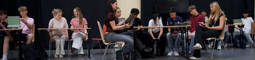 Eine Gruppe von Jugendlichen sitzt an Tischen in einem Raum. Einige arbeiten an Laptops, während andere auf Tablets oder Smartphones schauen. Die Atmosphäre wirkt konzentriert und die Jugendlichen scheinen an einer Aktivität oder einem Projekt beteiligt zu sein. Im Hintergrund sind dunkle Vorhänge zu sehen.