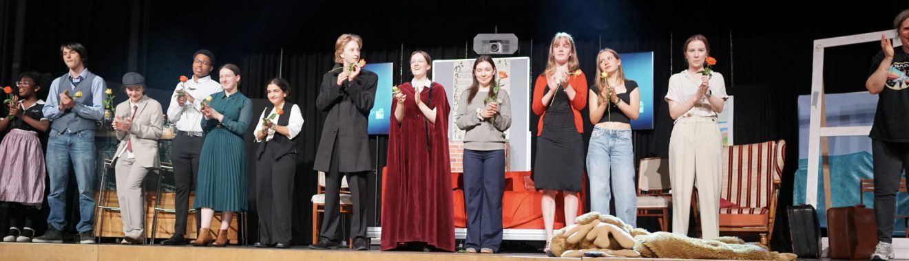 Eine Gruppe von Schauspielern steht auf einer Bühne und applaudiert. Sie tragen verschiedene Kostüme, die möglicherweise zu einem Theaterstück gehören. Im Hintergrund sind Bühnenbilder und Requisiten zu sehen. Die Atmosphäre wirkt festlich und begeistert.