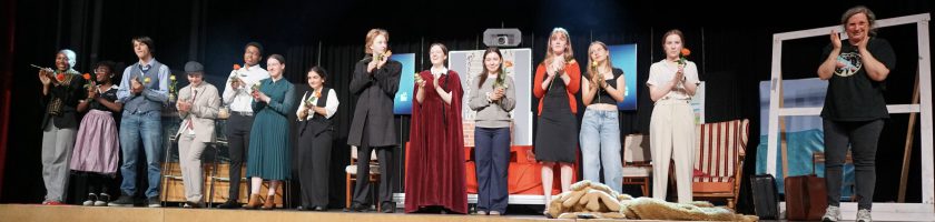 Eine Gruppe von Schauspielern steht auf einer Bühne und applaudiert. Sie tragen verschiedene Kostüme, die möglicherweise zu einem Theaterstück gehören. Im Hintergrund sind Bühnenbilder und Requisiten zu sehen. Die Atmosphäre wirkt festlich und begeistert.