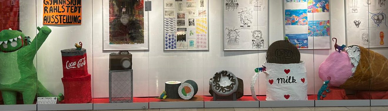 In einem Schaufenster sind mehrere künstlerische Objekte und Illustrationen ausgestellt. Auf der linken Seite steht eine große grüne Figur, die fröhlich wirkt. Daneben befinden sich verschiedene Behälter, darunter ein rotes mit dem Coca-Cola-Logo. In der Mitte liegt ein runder, dekorierter Tisch mit einem auffälligen Muster. Rechts steht eine große, braune Figur, die wie ein Eisbecher aussieht, und eine weitere Figur in rosa, die an einen großen, bunten Ballon erinnert. Im Hintergrund sind verschiedene Kunstwerke an der Wand zu sehen.