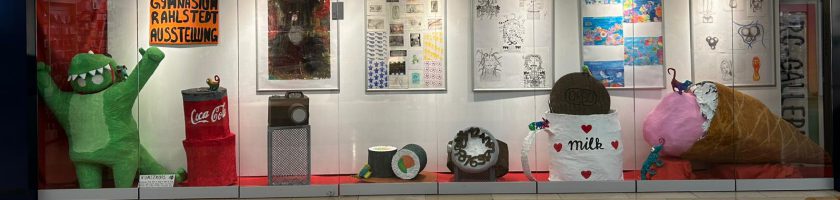 In einem Schaufenster sind mehrere künstlerische Objekte und Illustrationen ausgestellt. Auf der linken Seite steht eine große grüne Figur, die fröhlich wirkt. Daneben befinden sich verschiedene Behälter, darunter ein rotes mit dem Coca-Cola-Logo. In der Mitte liegt ein runder, dekorierter Tisch mit einem auffälligen Muster. Rechts steht eine große, braune Figur, die wie ein Eisbecher aussieht, und eine weitere Figur in rosa, die an einen großen, bunten Ballon erinnert. Im Hintergrund sind verschiedene Kunstwerke an der Wand zu sehen.