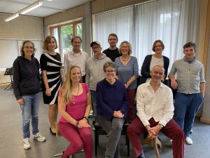 Eine Gruppe von zwölf Personen steht und sitzt in einem hellen Raum mit Fenstern und Vorhängen. Die Personen sind unterschiedlich gekleidet und lächeln in die Kamera. Im Vordergrund sitzen drei Personen, während die anderen im Hintergrund stehen. Der Raum hat einen neutralen Boden und eine schlichte Einrichtung.
